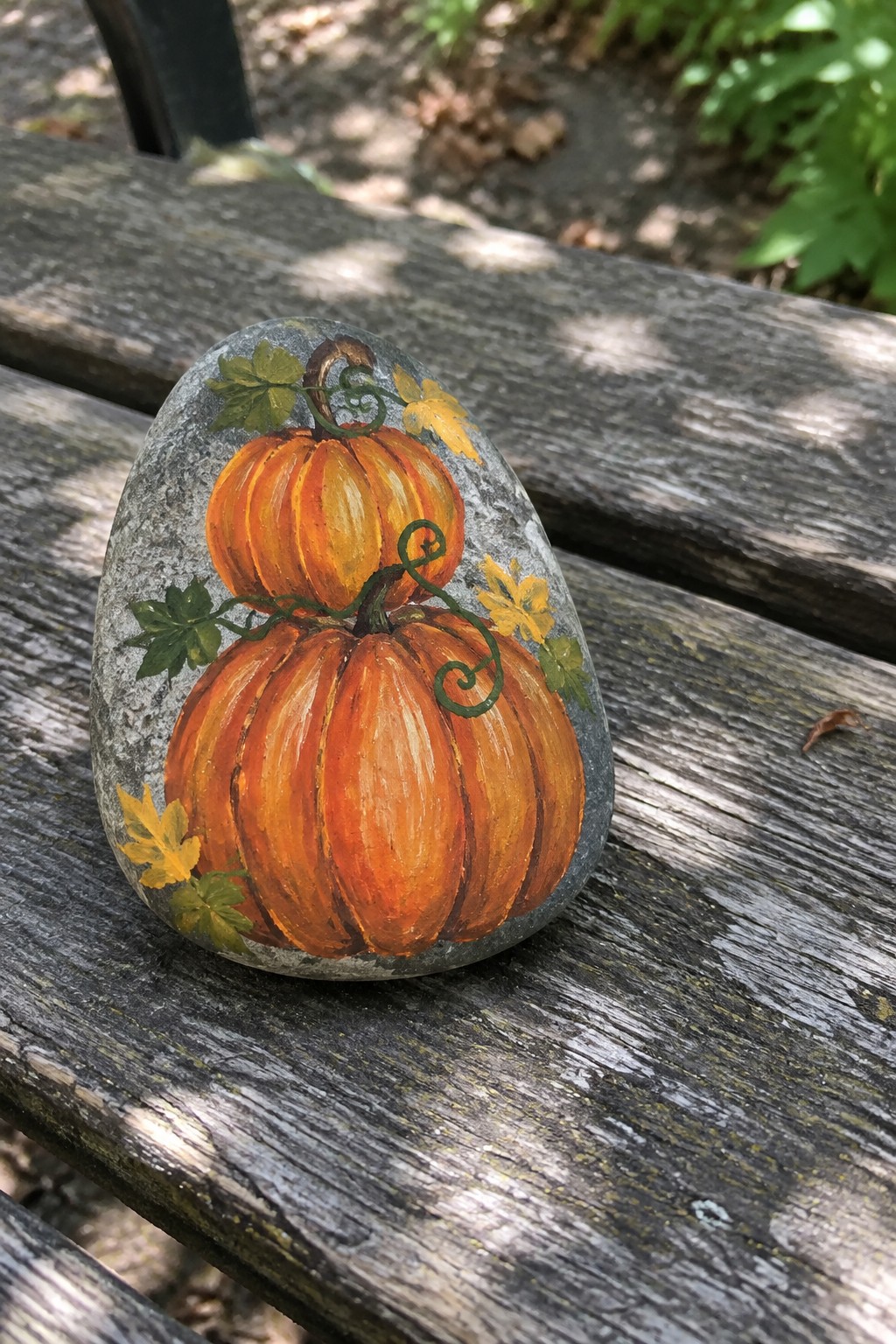 Painted gray rock with two orange pumpkins, green vines, and yellow leaves on wood.
