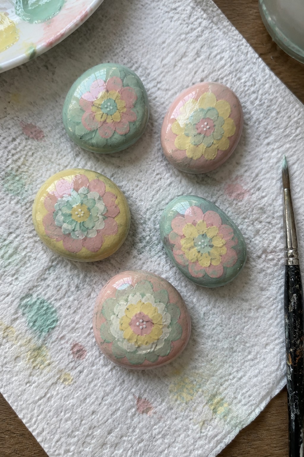 Five glossy pastel floral painted rocks on paper towel beside paintbrush