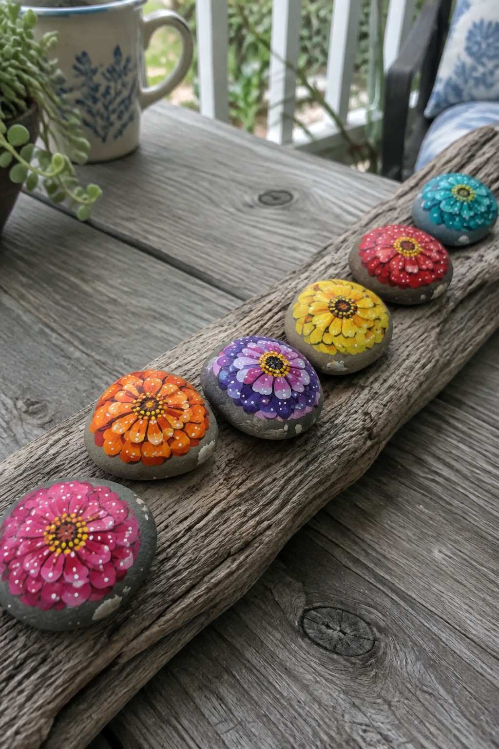 Six colorful painted rocks with zinnia flowers on weathered driftwood.
