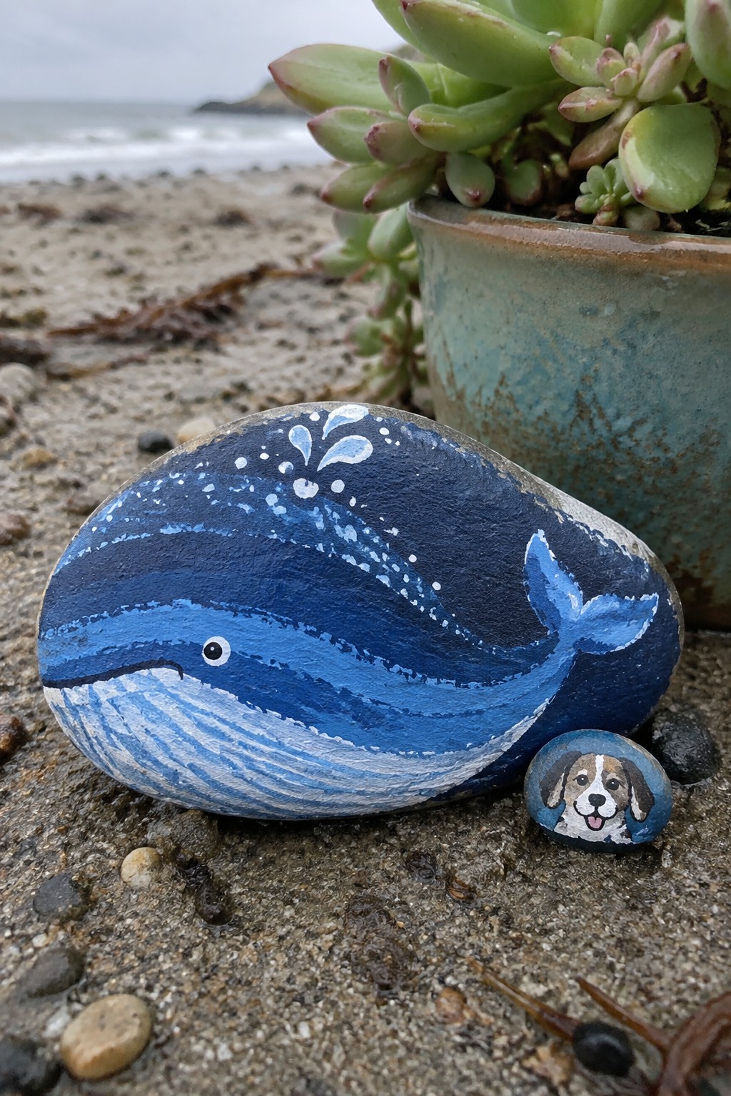 Blue whale rock painting beside small dog-faced pebble on beach near potted succulent