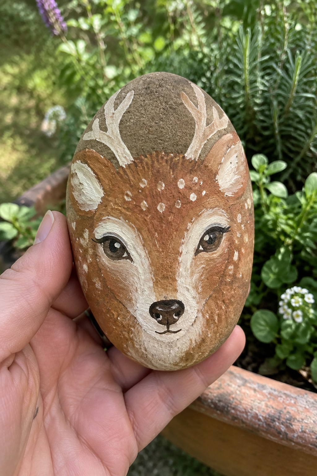 Hand holds oval rock painted as deer face with antlers, brown fur, white spots