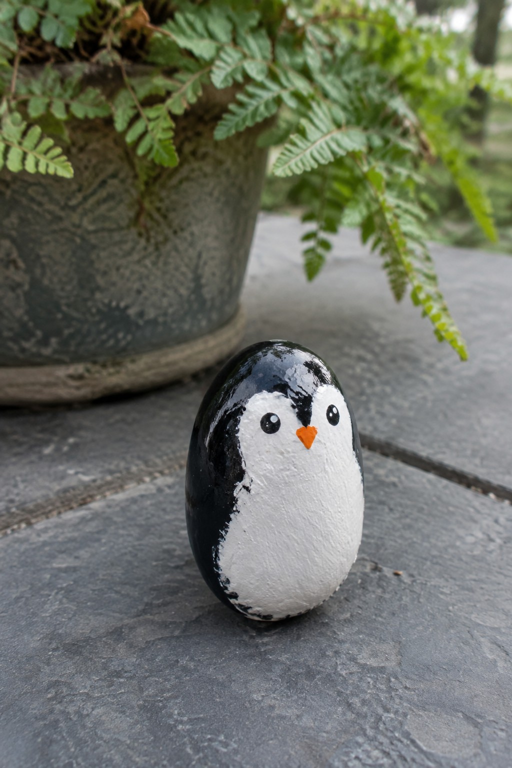 Black and white painted rock penguin with orange beak on stone near potted plant