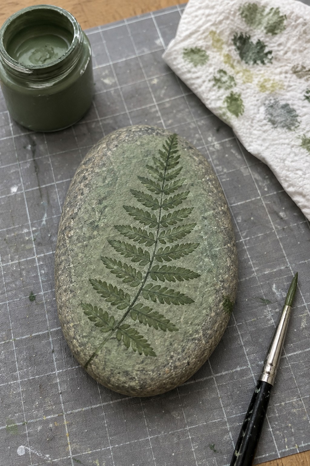 Stone with green fern print beside paint jar, brush and stamped paper towel on mat.
