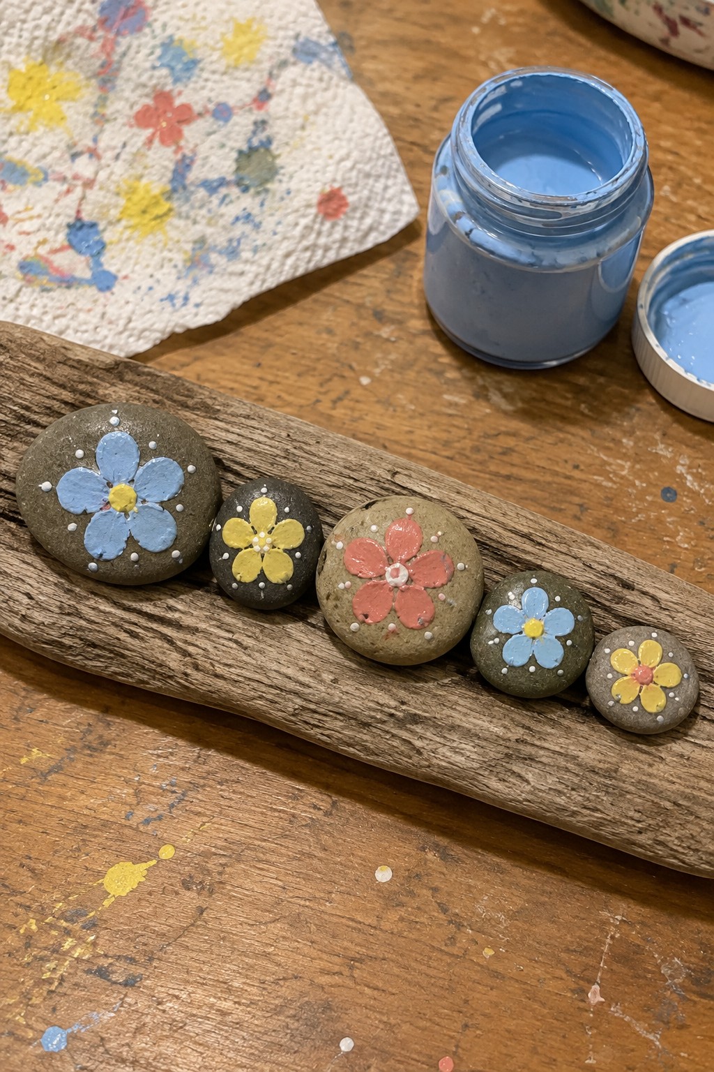 Five flower-painted rocks lined on driftwood beside open blue paint jar and splattered towel