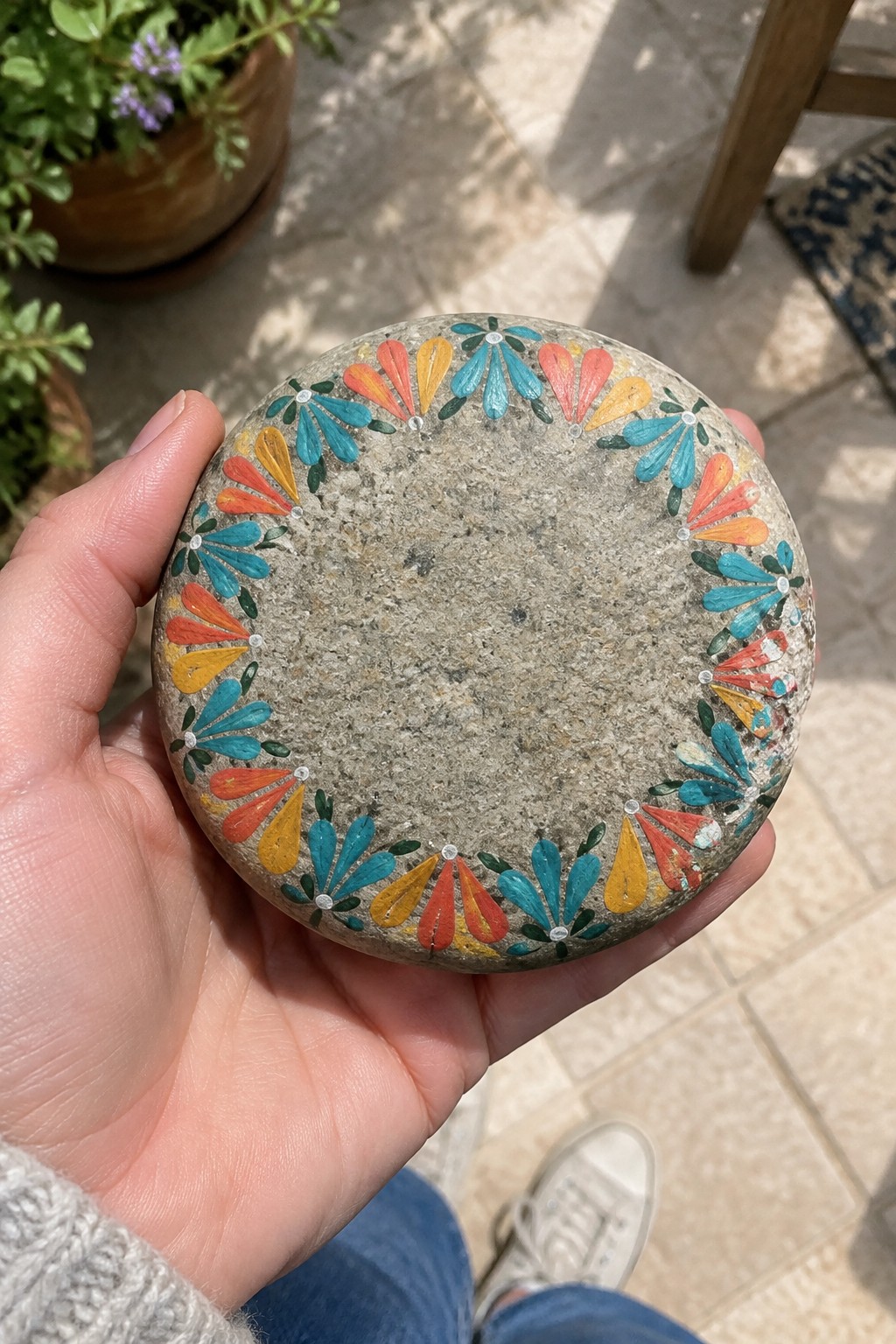 Hand holding round stone with colorful fan-shaped painted border on patio.