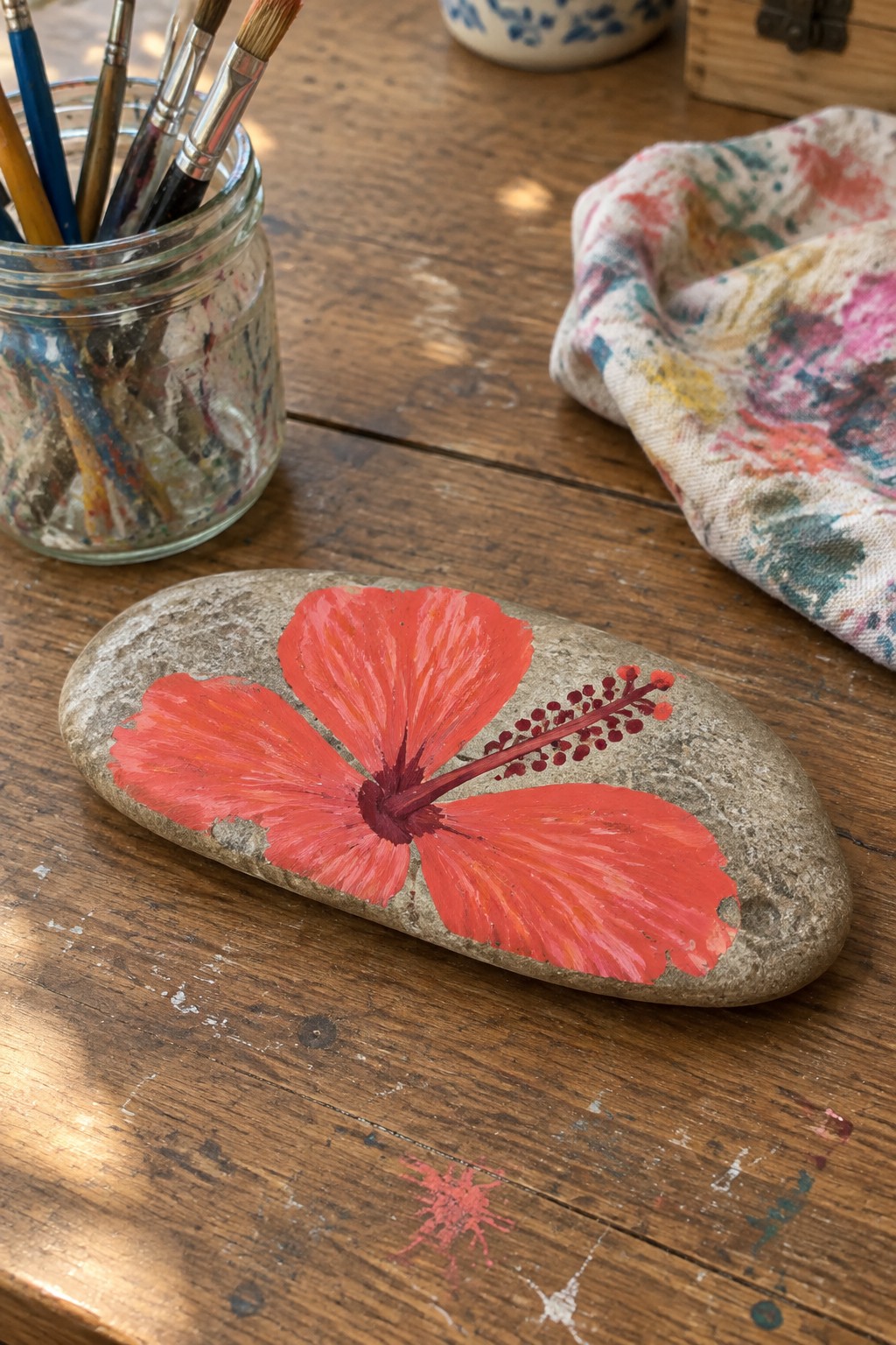 Oval stone with vibrant red hibiscus flower on paint-splattered wooden table beside brushes