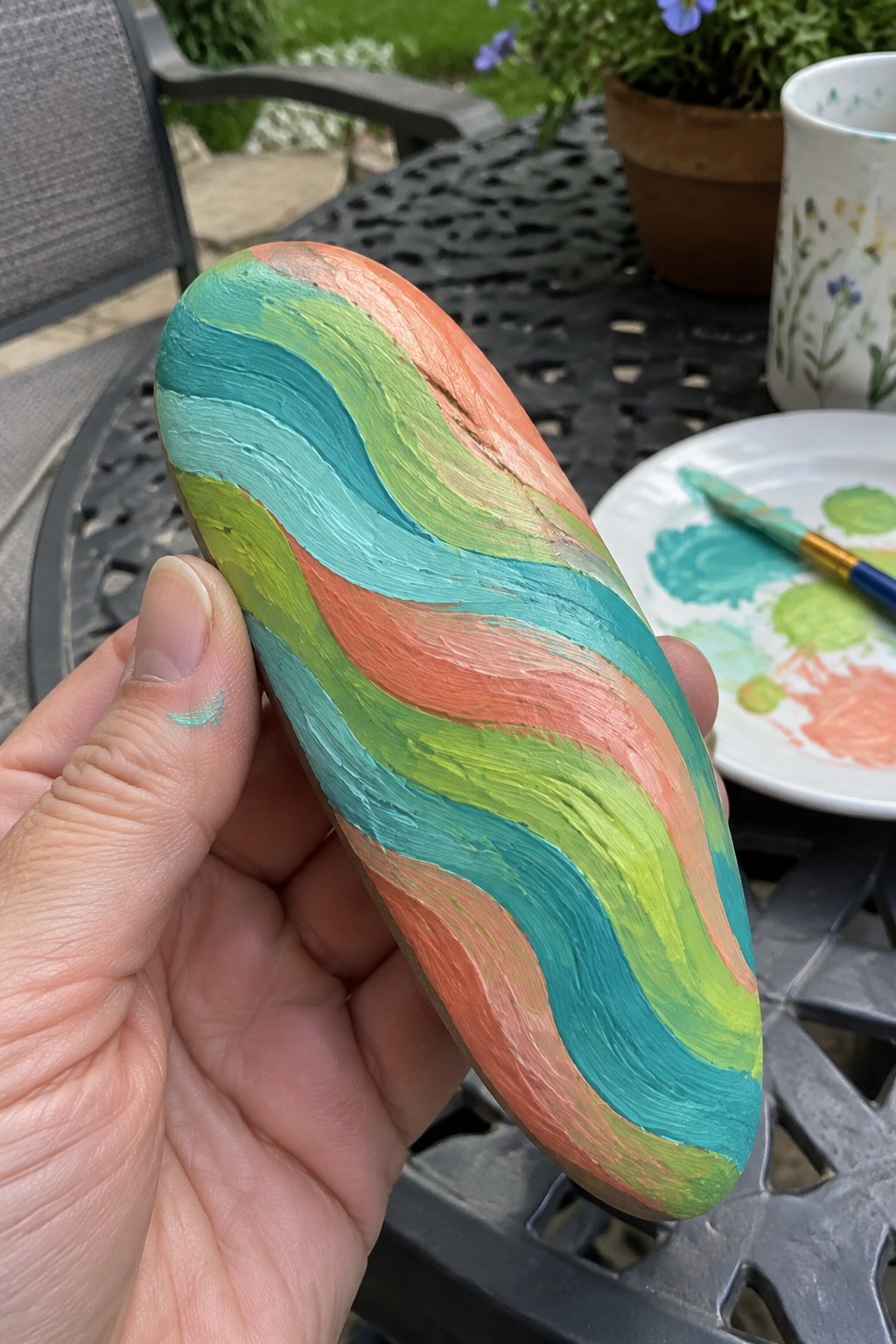 Hand holding oblong stone with wavy coral, teal, lime, and turquoise paint stripes.