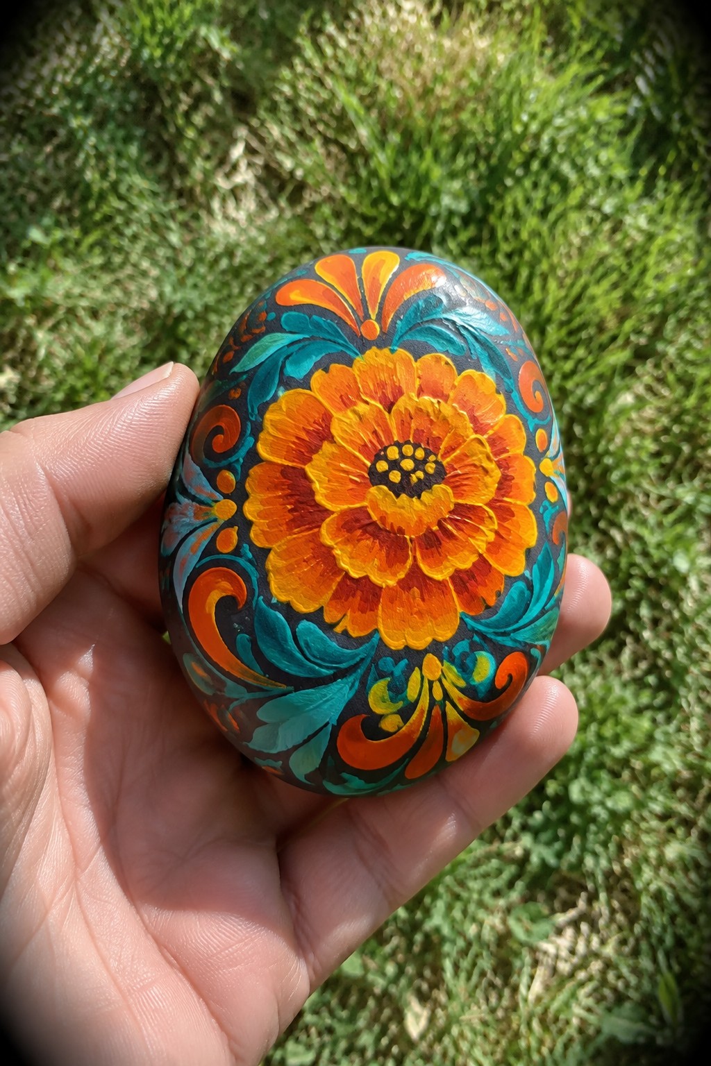 Hand holding egg-shaped stone painted with orange flower and teal swirls