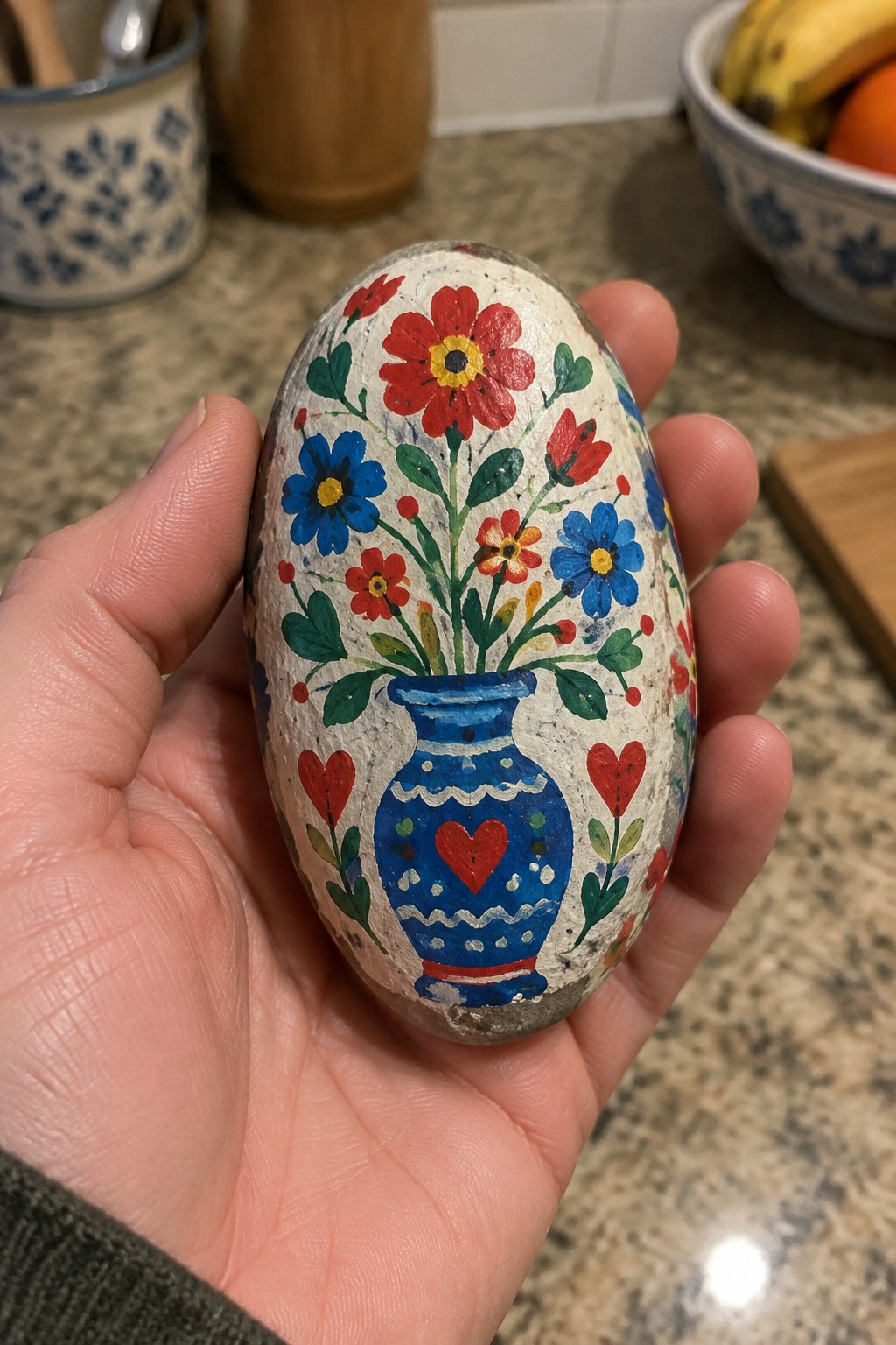 Hand holding oval painted rock with blue vase, colorful flowers, and red hearts.