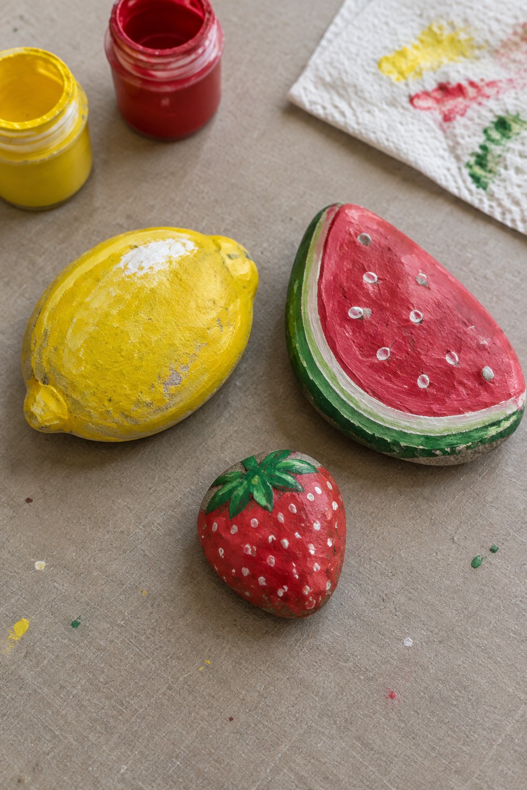 Three painted fruit rocks—a lemon, watermelon slice, and strawberry—beside paint jars