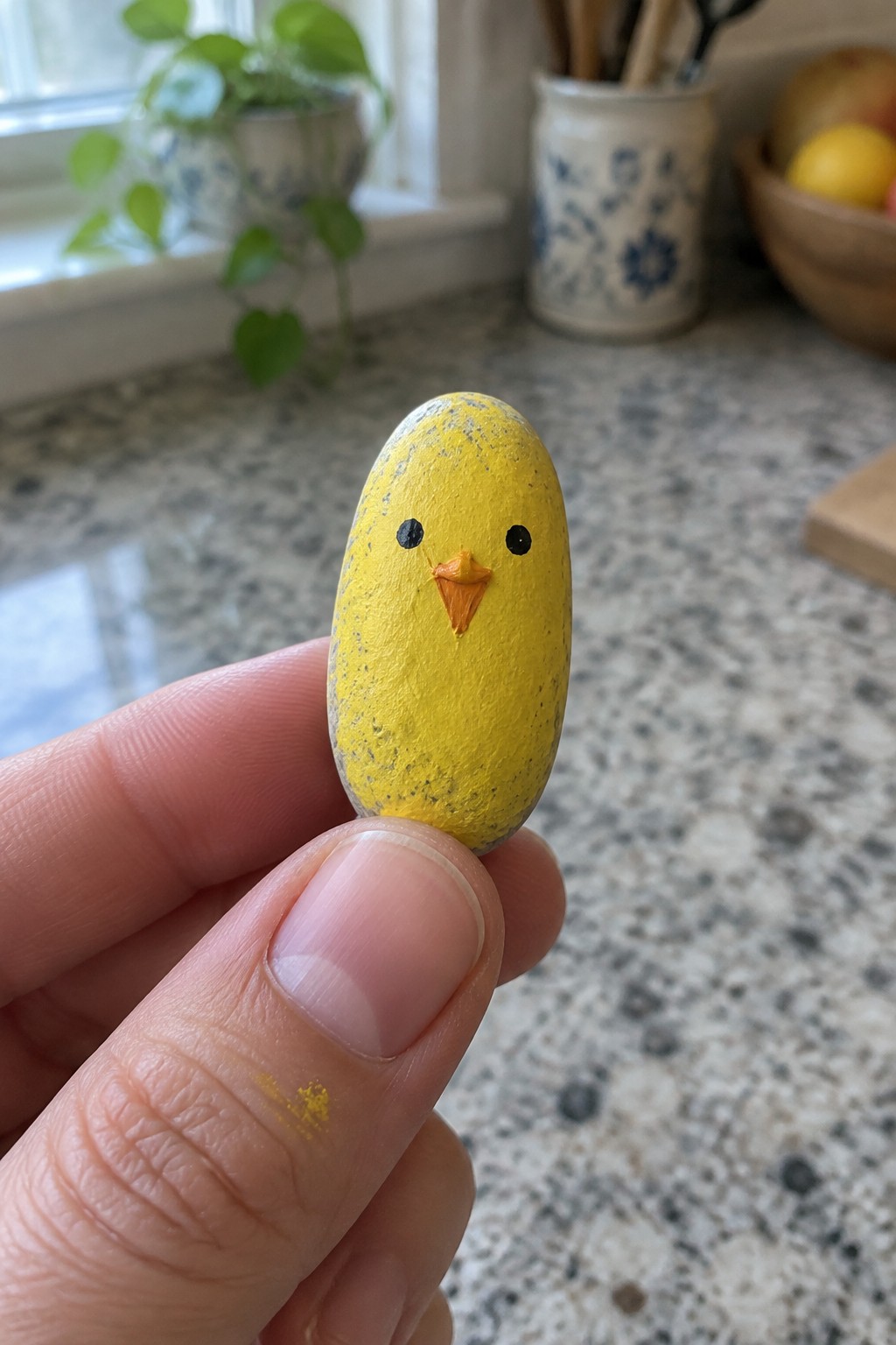Hand holding small yellow painted rock as chick with black eyes and orange beak