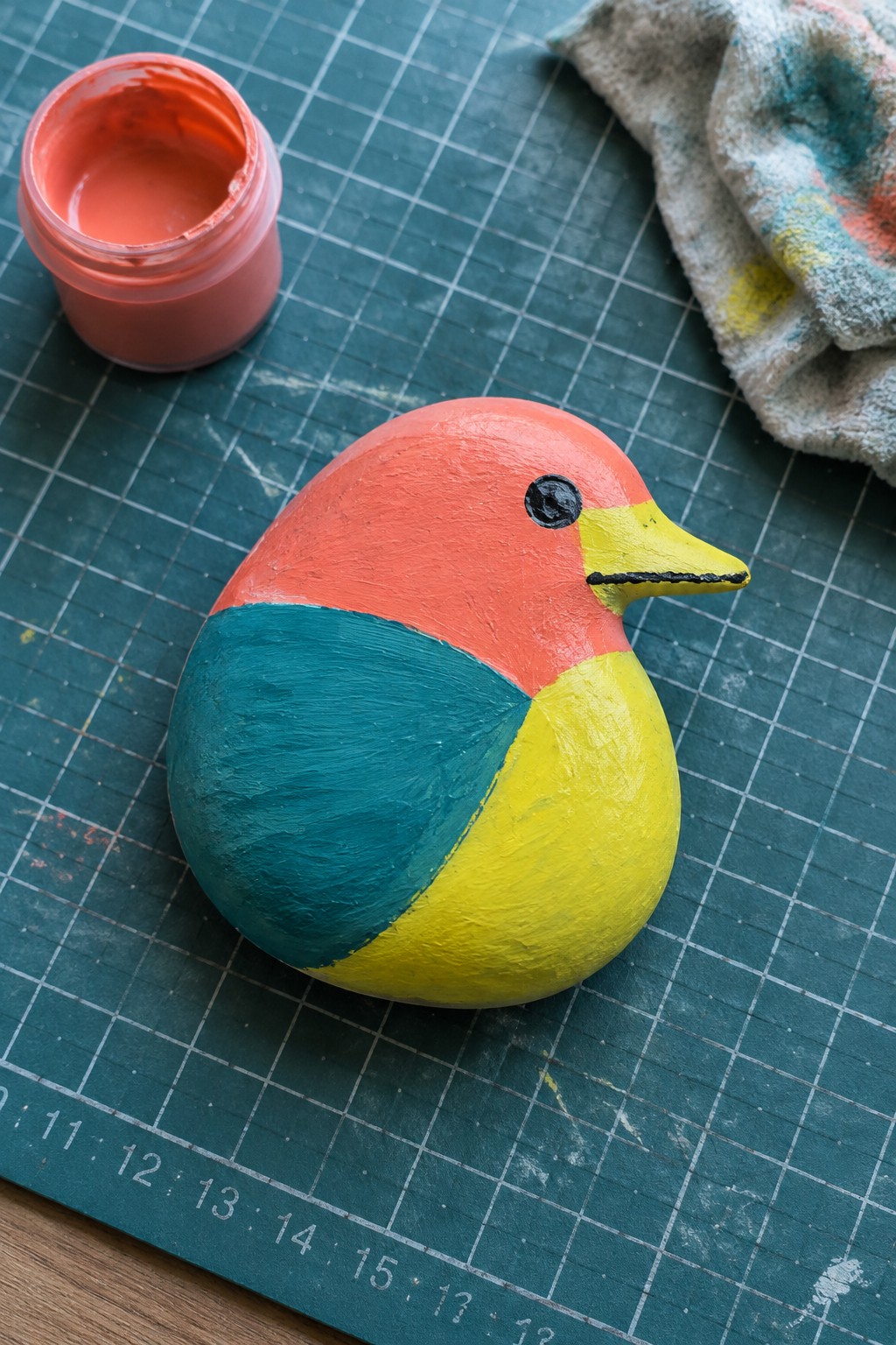 Red, teal, and yellow painted bird beside open paint jar and rag on cutting mat.