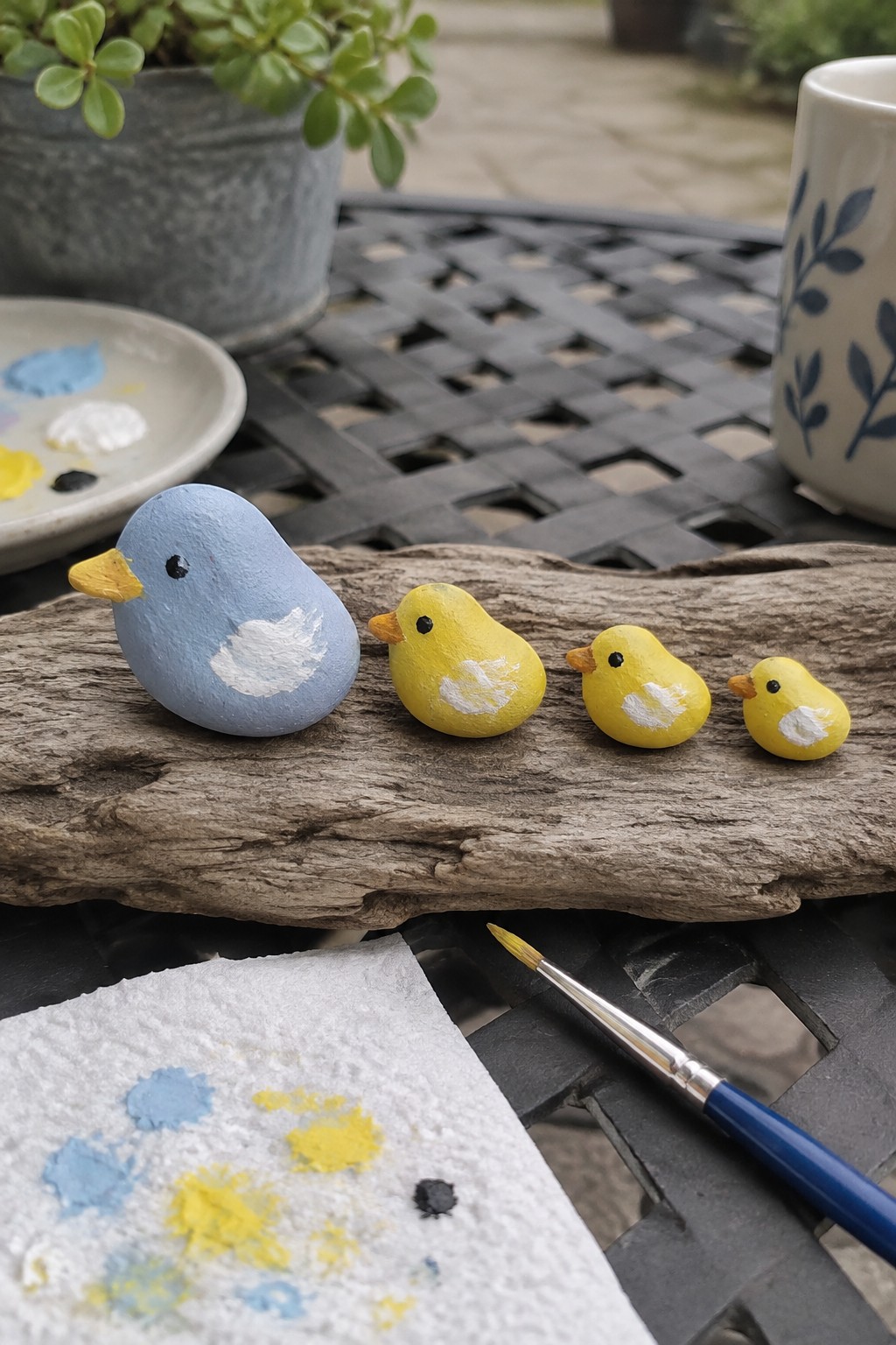 Painted blue and yellow rock birds of decreasing sizes on weathered driftwood with supplies.