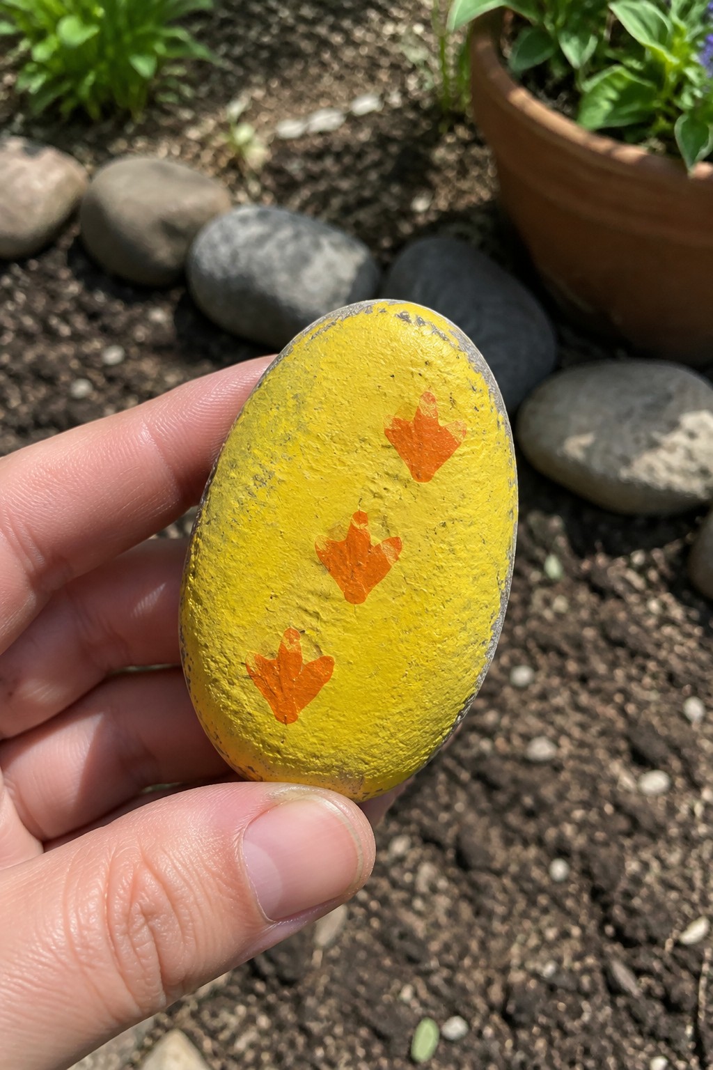 Hand holding yellow painted rock with three orange crown symbols in garden