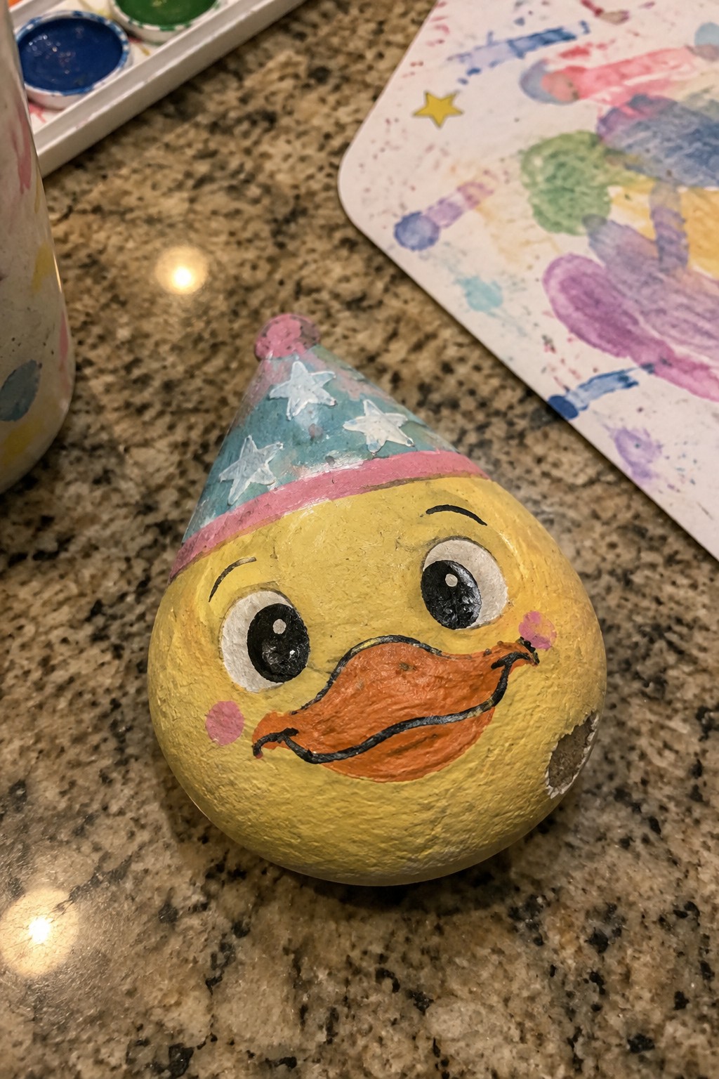 Painted yellow duck rock with orange beak wearing blue starry party hat on granite
