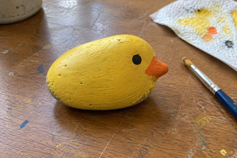 19 Easy Duck Rock Painting Ideas With Recognizable Shapes Yellow painted rock chick with orange beak and black eye on wooden table