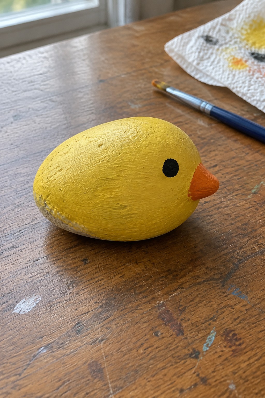 Yellow painted rock chick with orange beak and black eye on wooden table