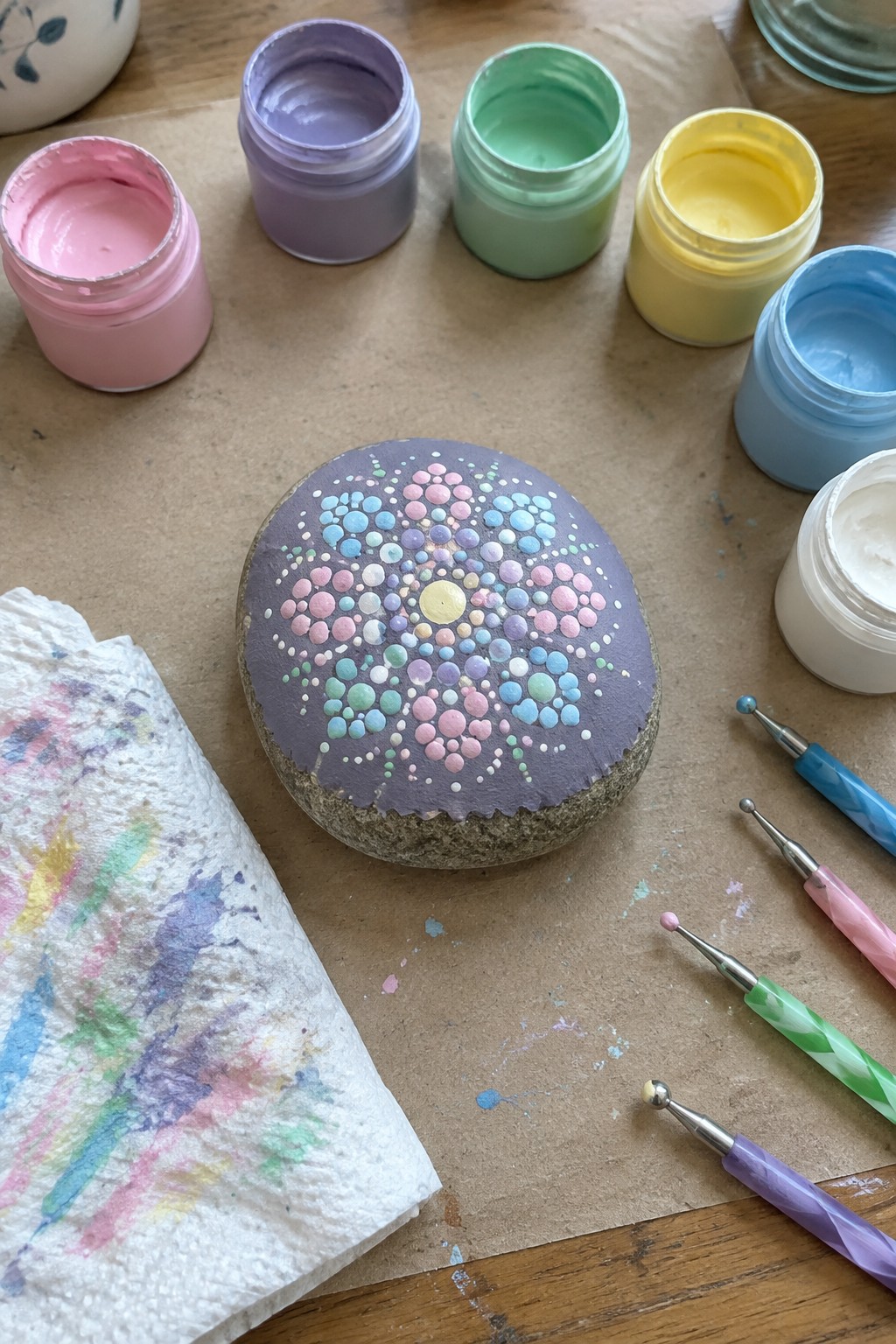 Painted rock with colorful dot mandala, paint jars, and dotting tools on table