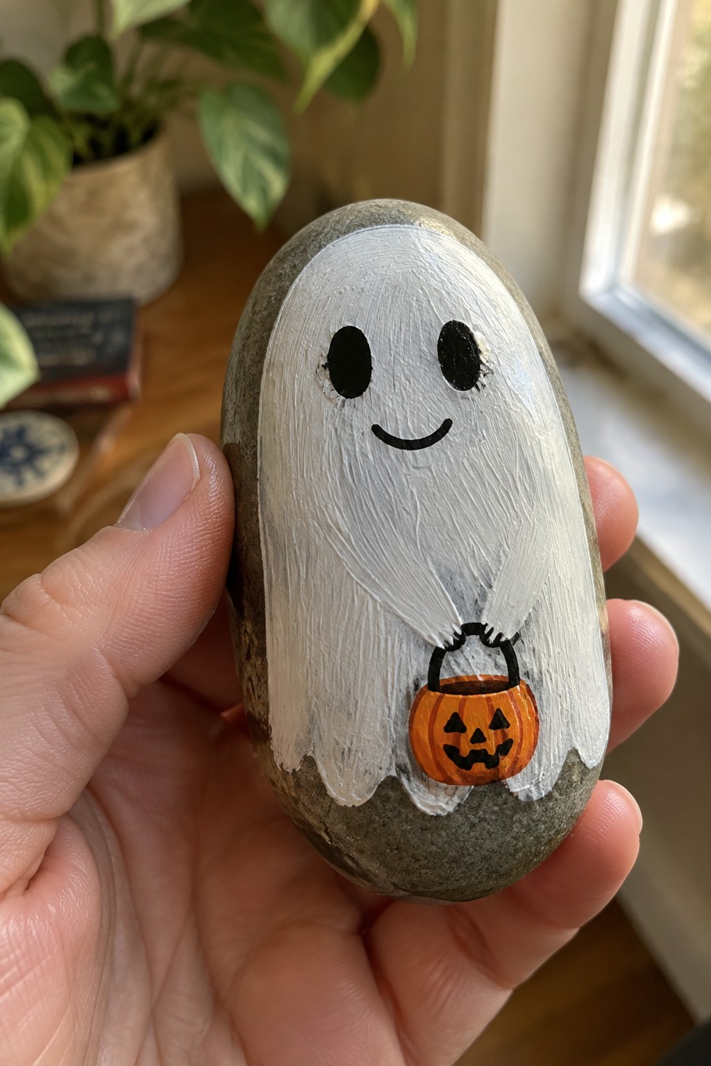 Hand holding oval rock painted as white ghost with black eyes clutching pumpkin bucket