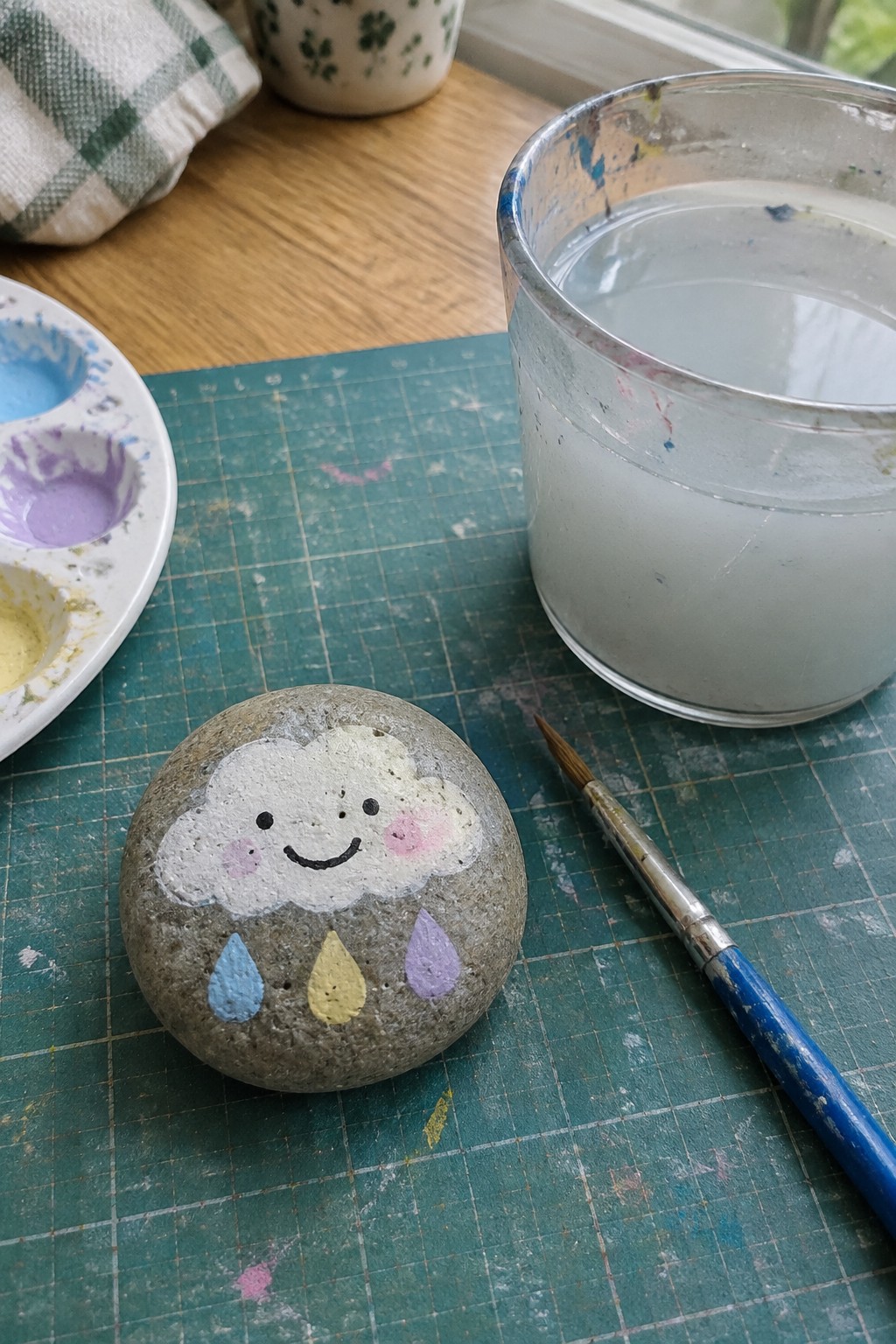 Painted rock with smiling cloud and blue-yellow-purple raindrops beside paint palette, brush, and water glass