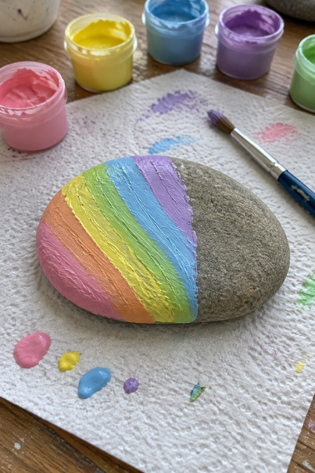 Rainbow-striped painted rock beside paint jars, brush, and dabs on paper towel.