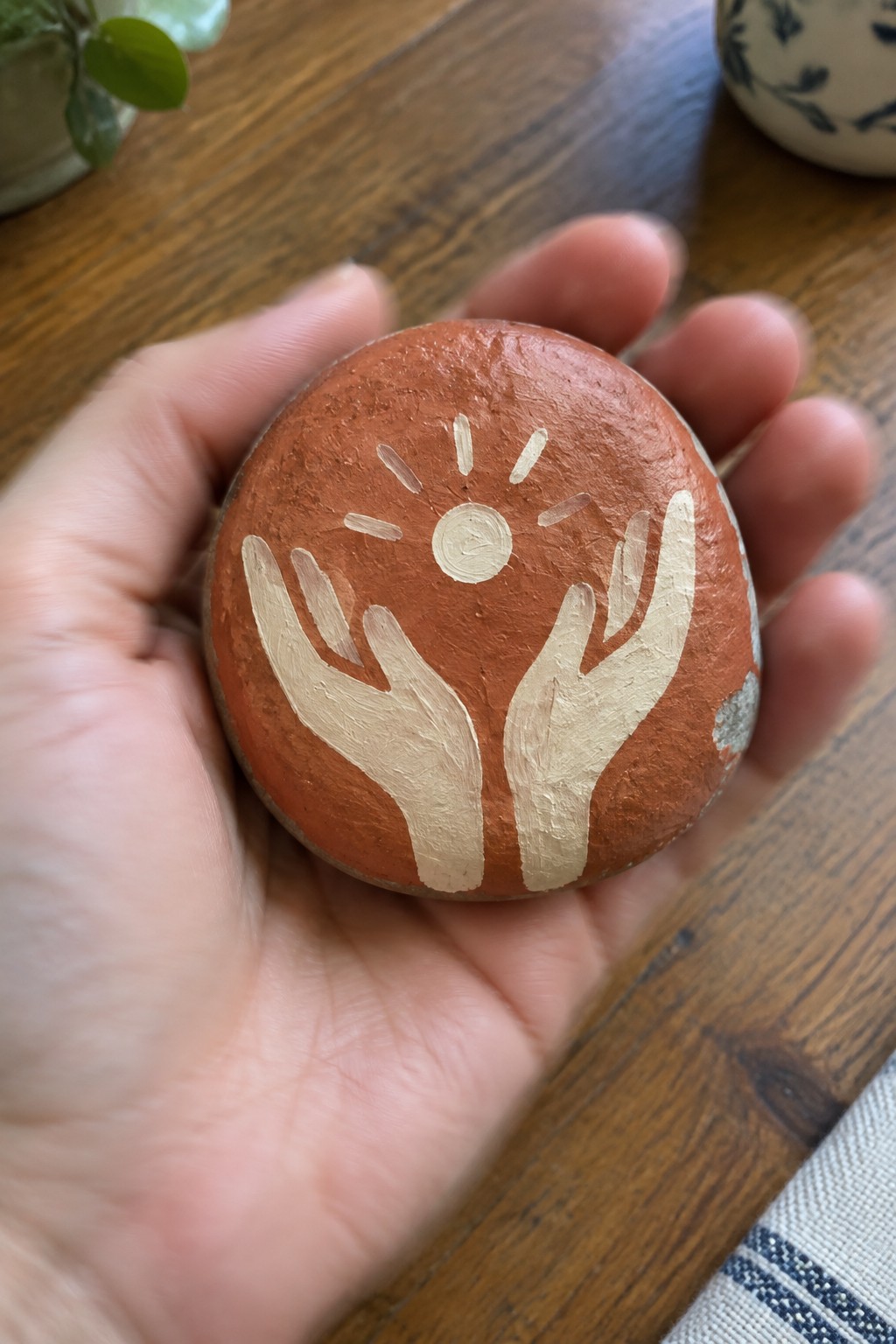 A hand holds a round reddish-brown painted rock showing two light-colored hands reaching up toward a small sun with rays.