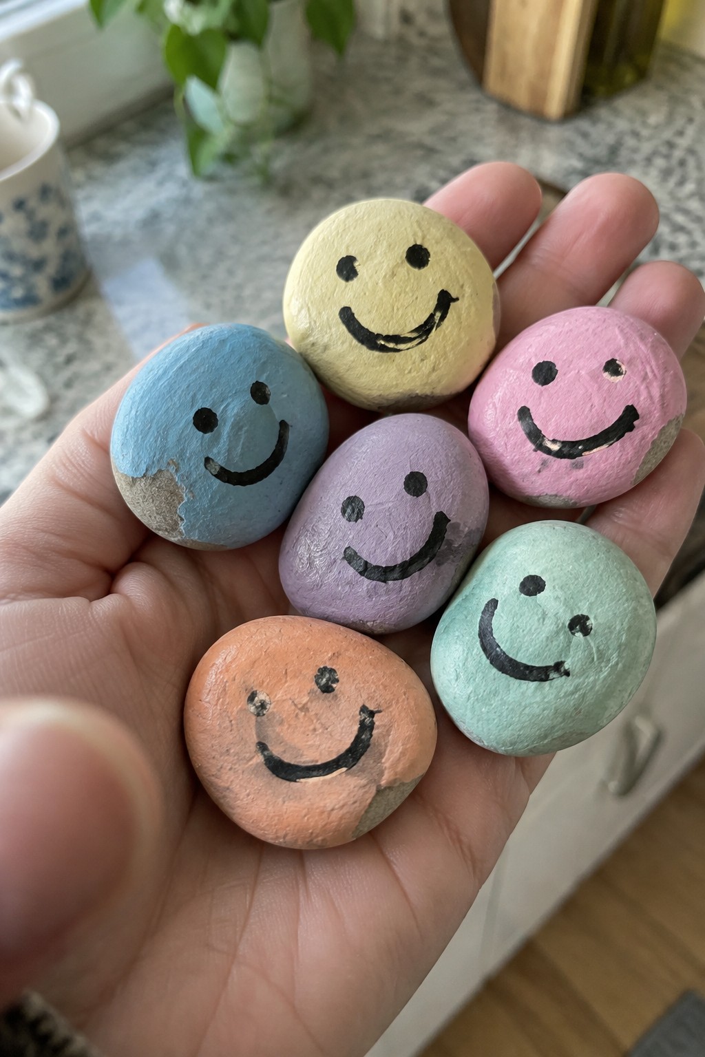 Six small smooth rocks painted in pastel colors with black smiley faces held in a person's hand.