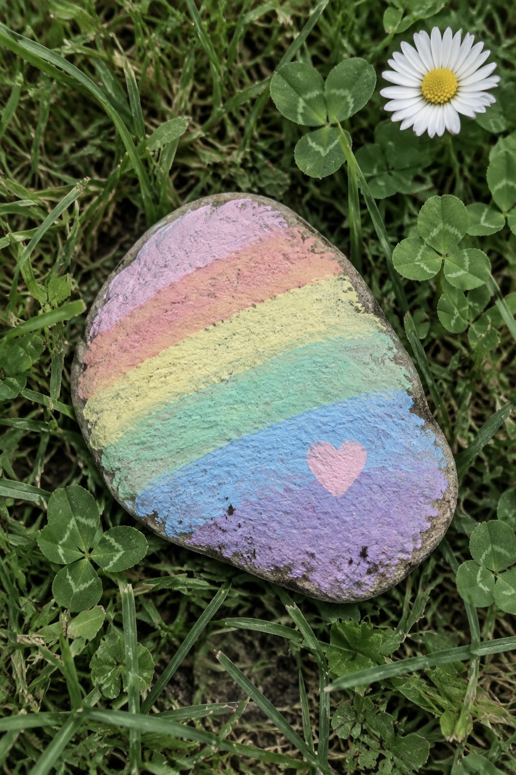 Rainbow-striped rock with pink heart on grass near clover and daisy
