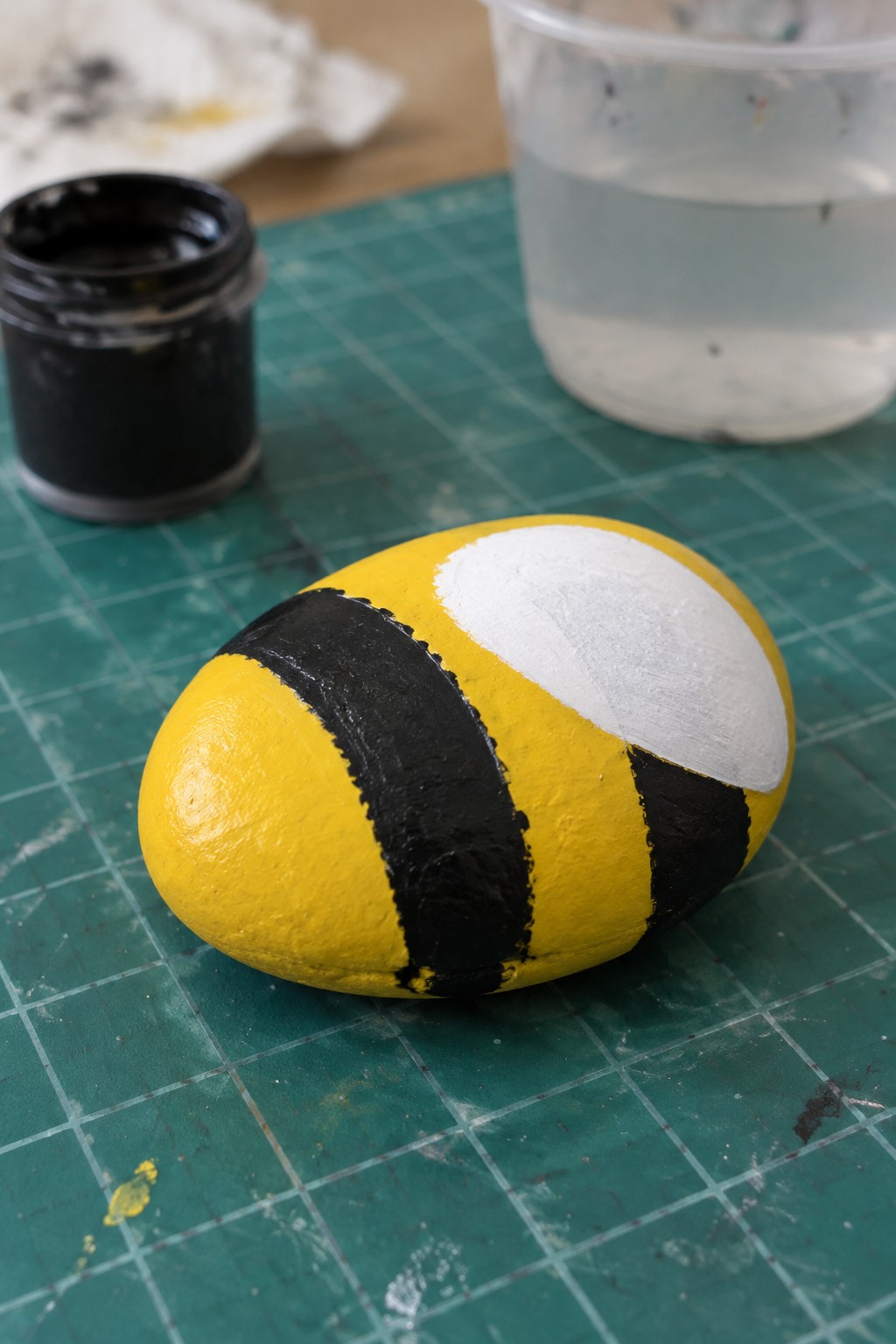 Yellow black striped painted rock on green grid mat with paint jar