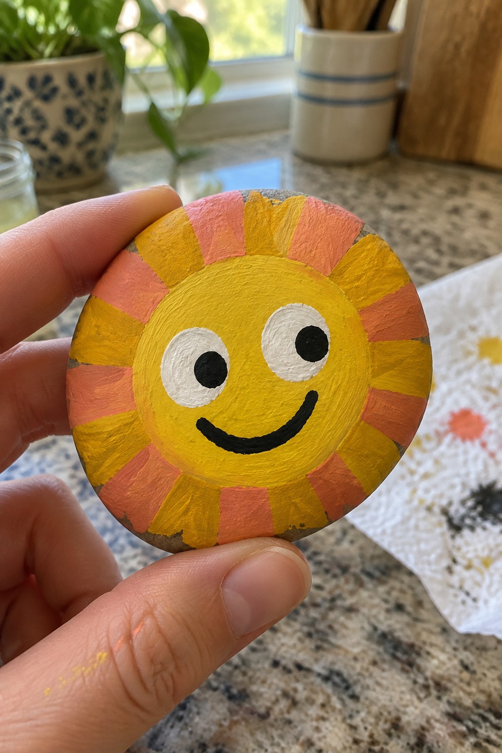 Hand holding a smiling sun painted on a rock with yellow-orange rays.
