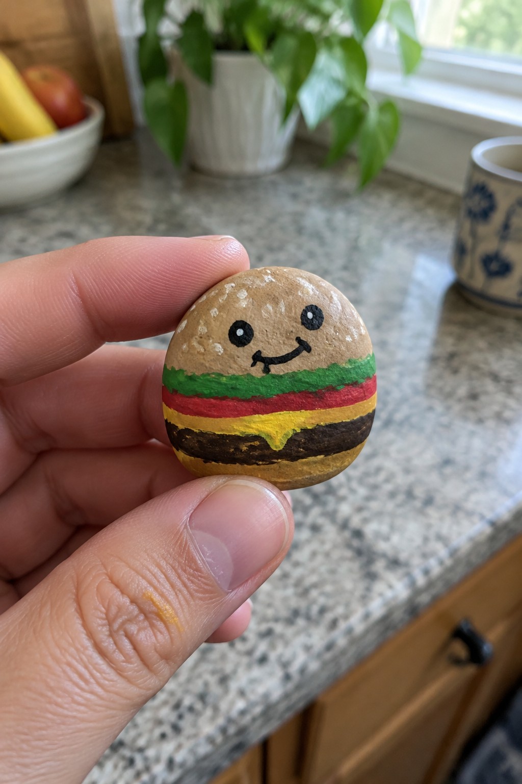 Fingers holding smiling hamburger rock painted with bun, lettuce, tomato, cheese, and patty.