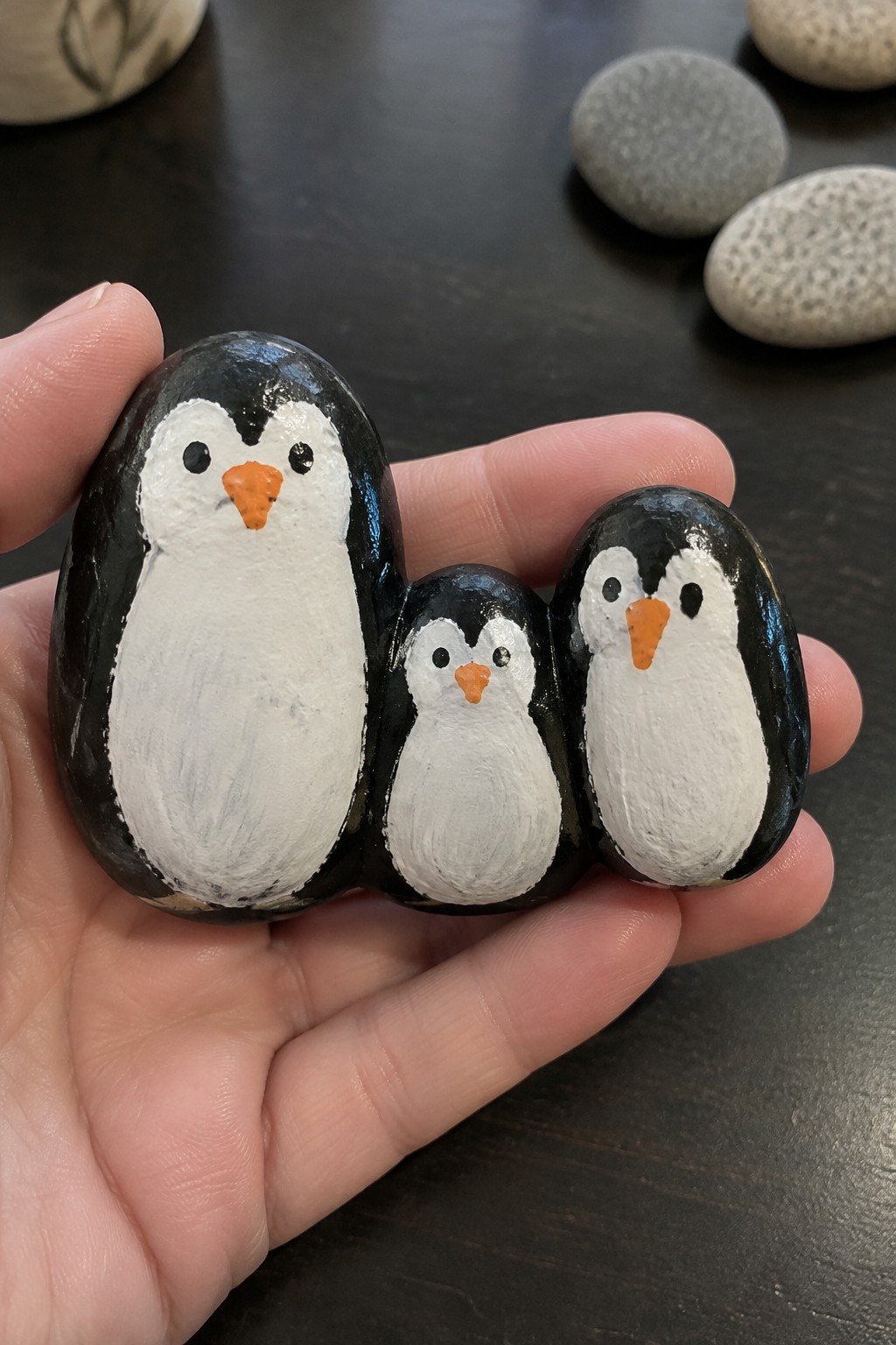 Hand holding three black-and-white painted penguin rocks with orange beaks.