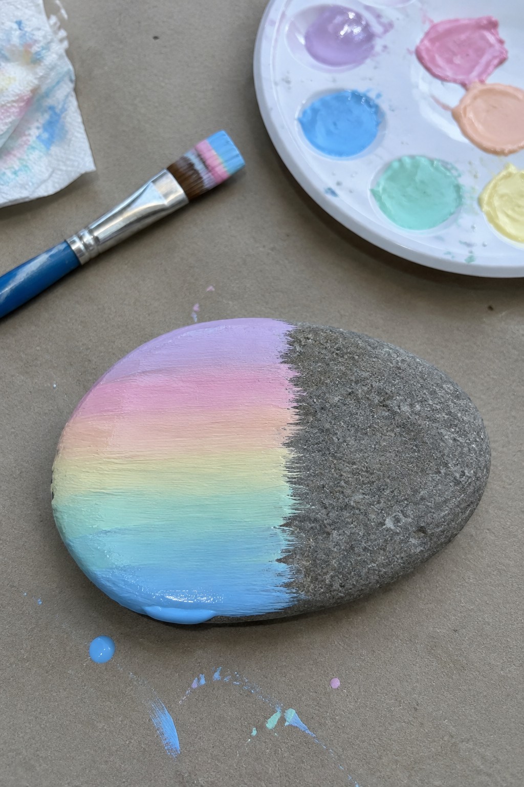 Rainbow-painted rock half-covered in pastel stripes beside palette and brush