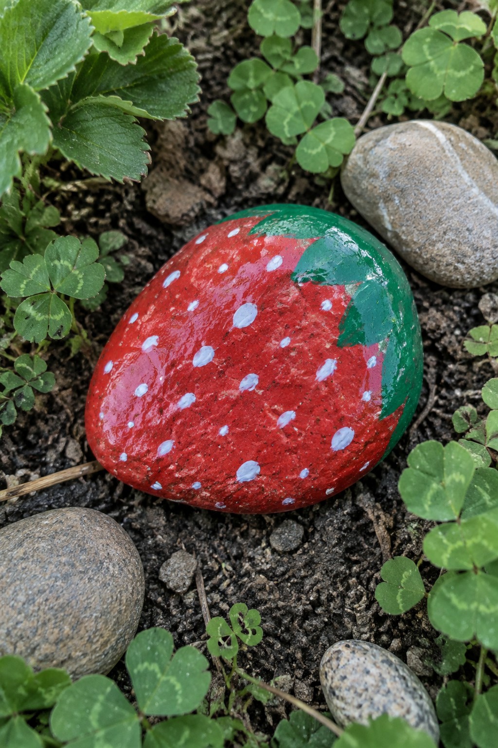Red strawberry rock painted with white dots and green leaves amid soil and plants.