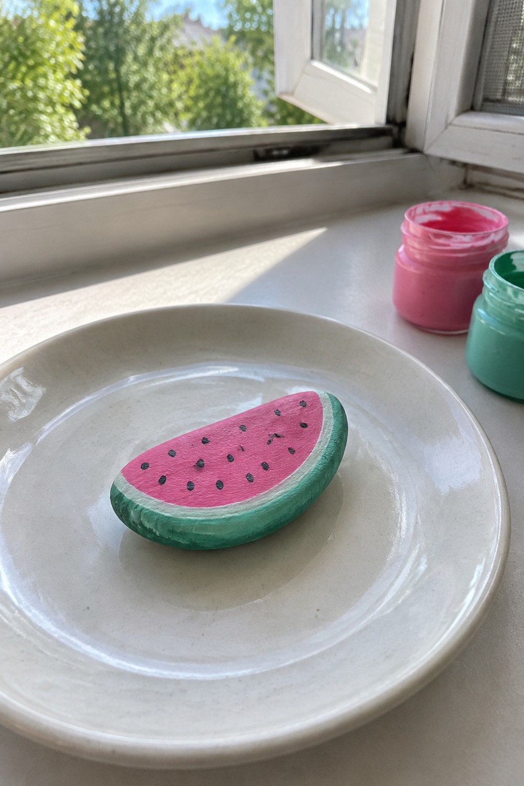 Watermelon-painted rock on white plate beside pink and green paint jars on windowsill.
