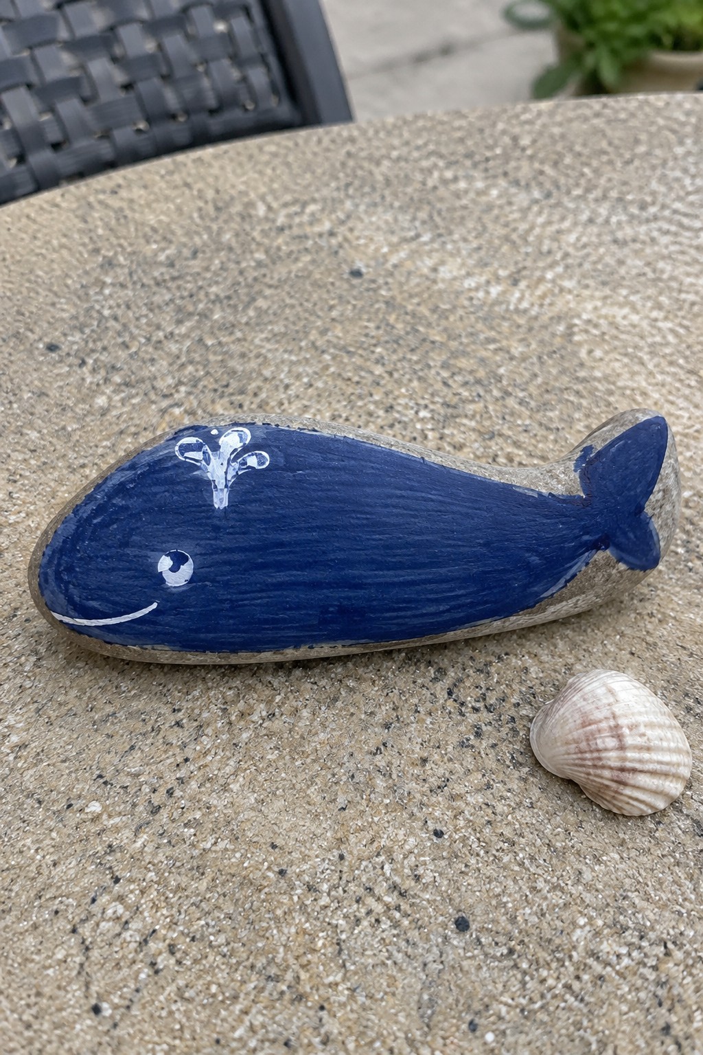A blue painted whale on an elongated rock sits on a stone table next to a small seashell.