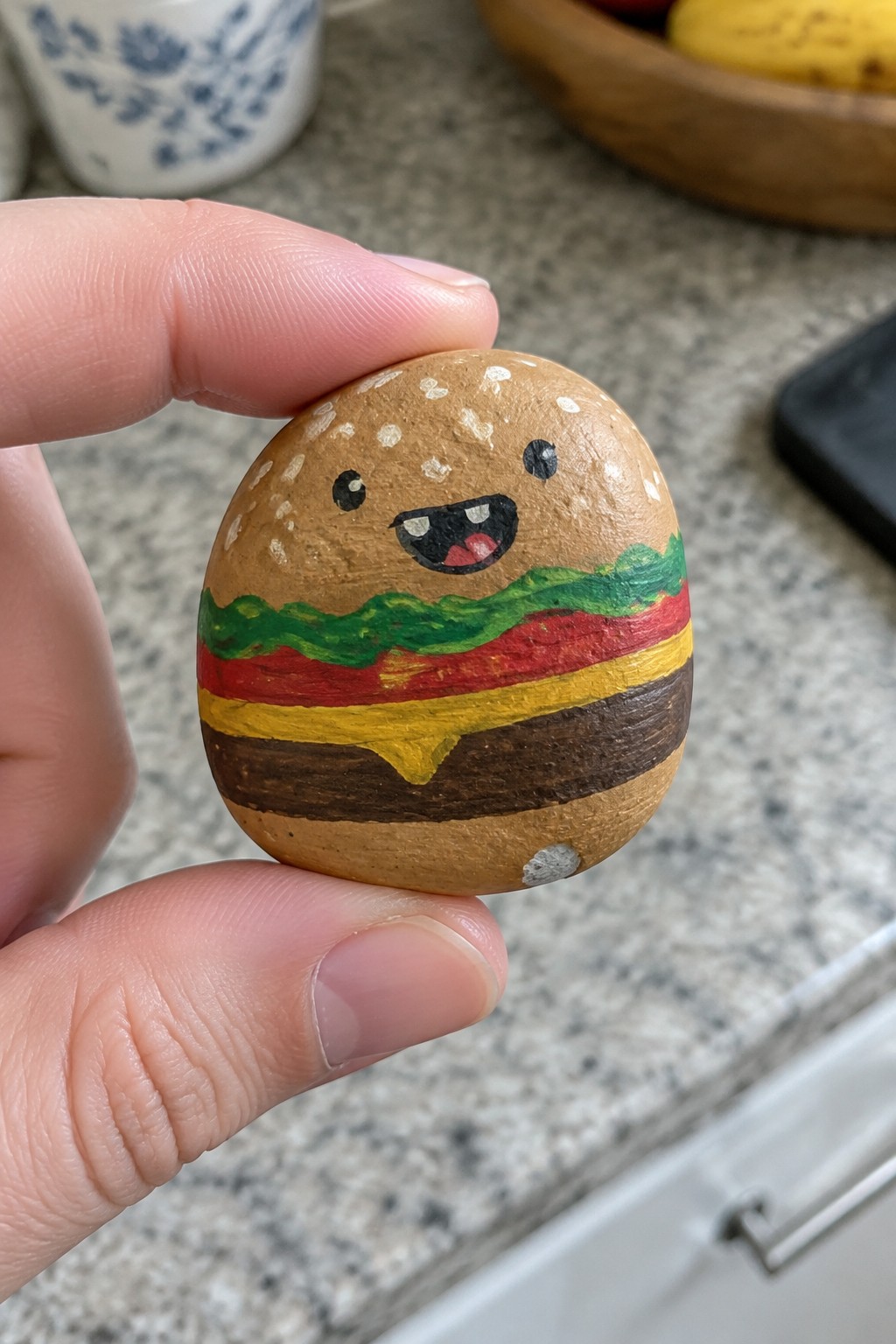 A hand holds a small rounded rock painted like a hamburger with a cute face and layered colors for bun, lettuce, tomato, cheese, and patty.