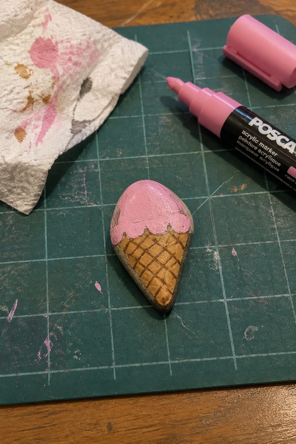 Painted rock ice cream cone with pink scoop beside uncapped Posca marker on green mat
