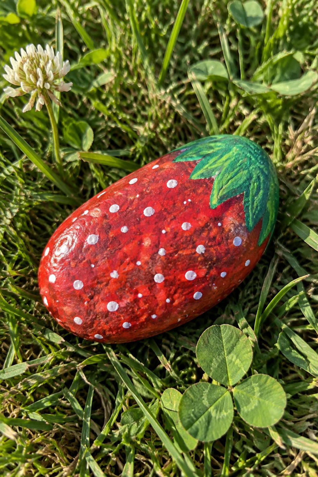 Red strawberry painted rock with white dots and green leaves on grass