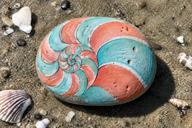 18 Summer Rock Painting Ideas Inspired by Waves and Seashells Orange and teal nautilus spiral painted on rock amid shells on wet sand