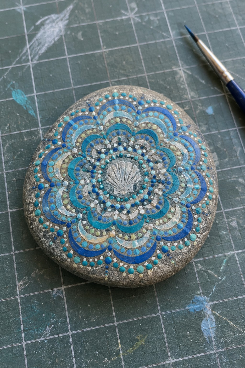Painted stone with blue mandala dots, arcs, and central seashell beside paintbrush