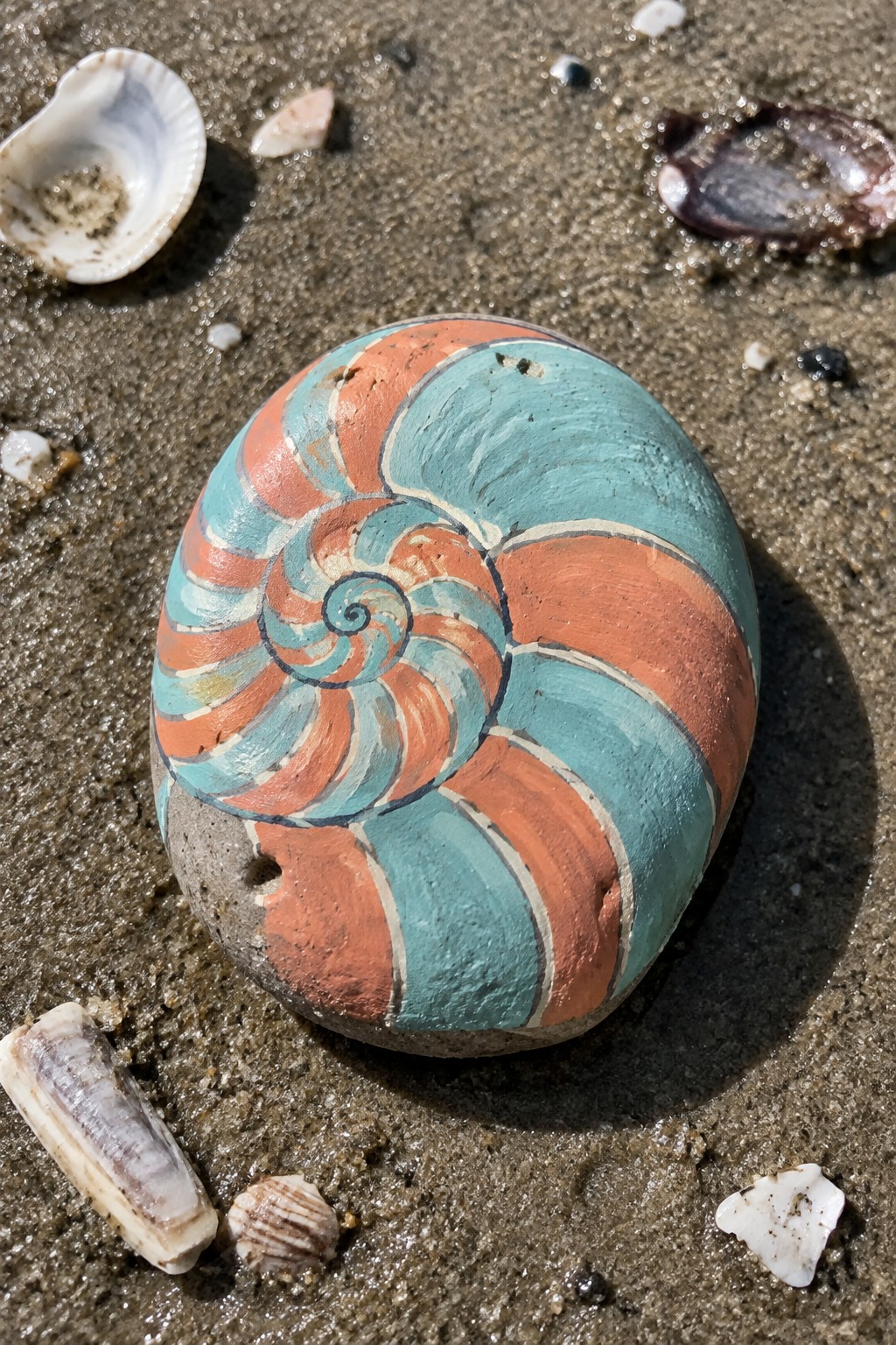 Orange and teal nautilus spiral painted on rock amid shells on wet sand