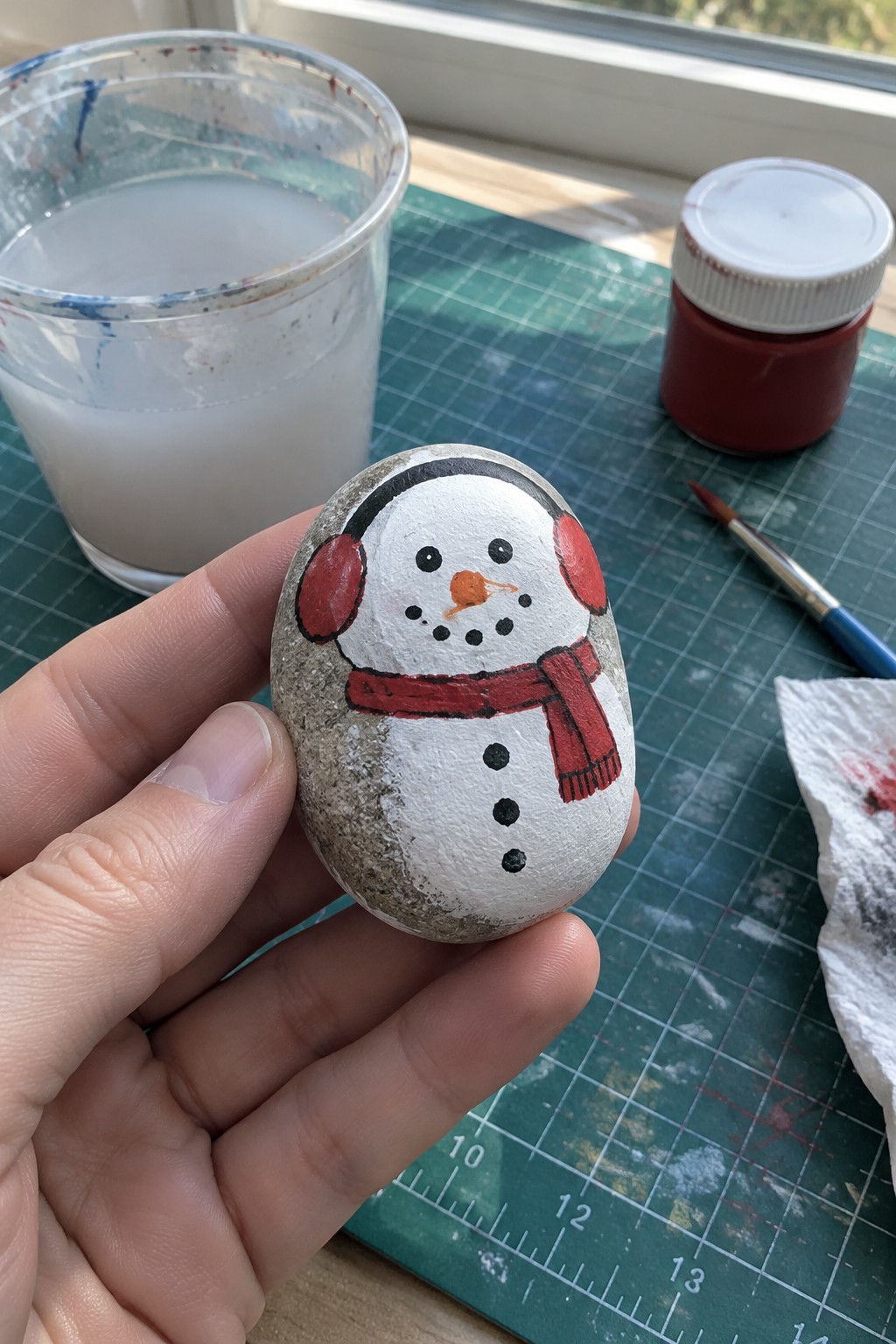 Hand holding painted stone snowman with red scarf, earmuffs, and buttons on green mat