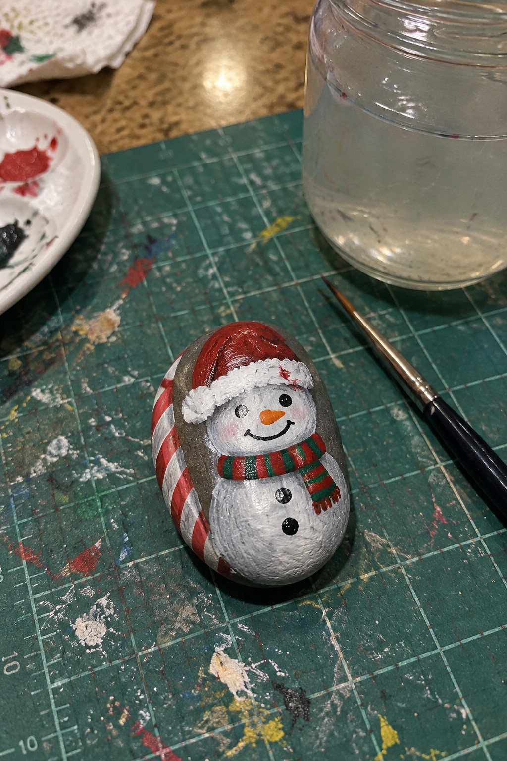 Painted rock snowman with red Santa hat, striped scarf on splattered green mat with supplies