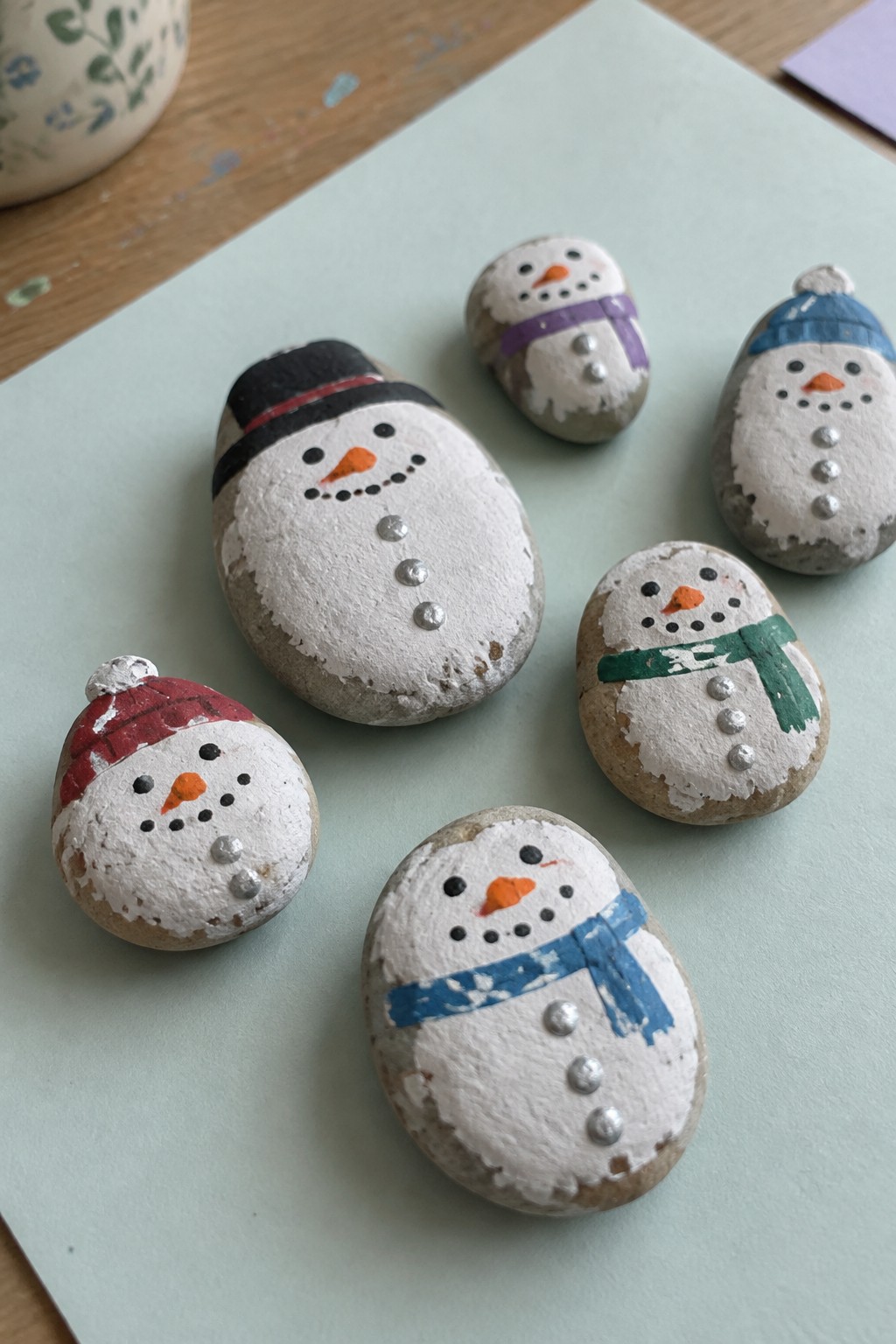 Five painted rock snowmen with colorful hats and scarves on green paper