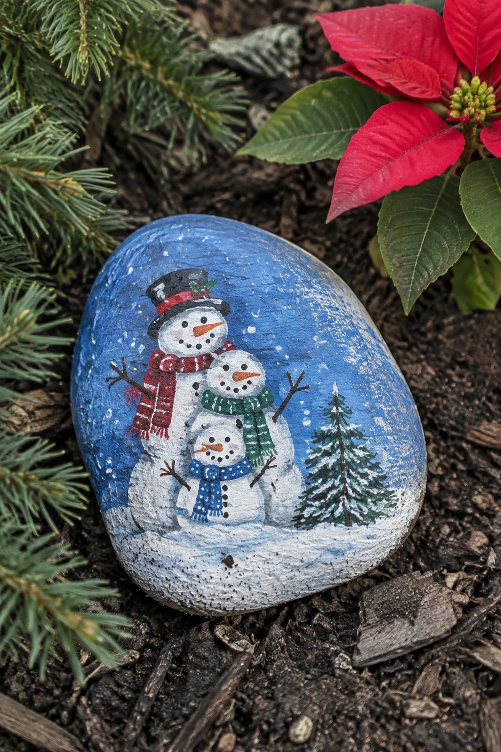 Three snowmen with colorful scarves painted on a rock beside poinsettia and pine.