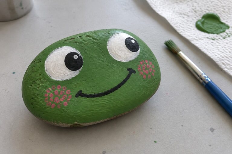 Green painted rock frog with white eyes, pink cheeks, black smile beside paintbrush