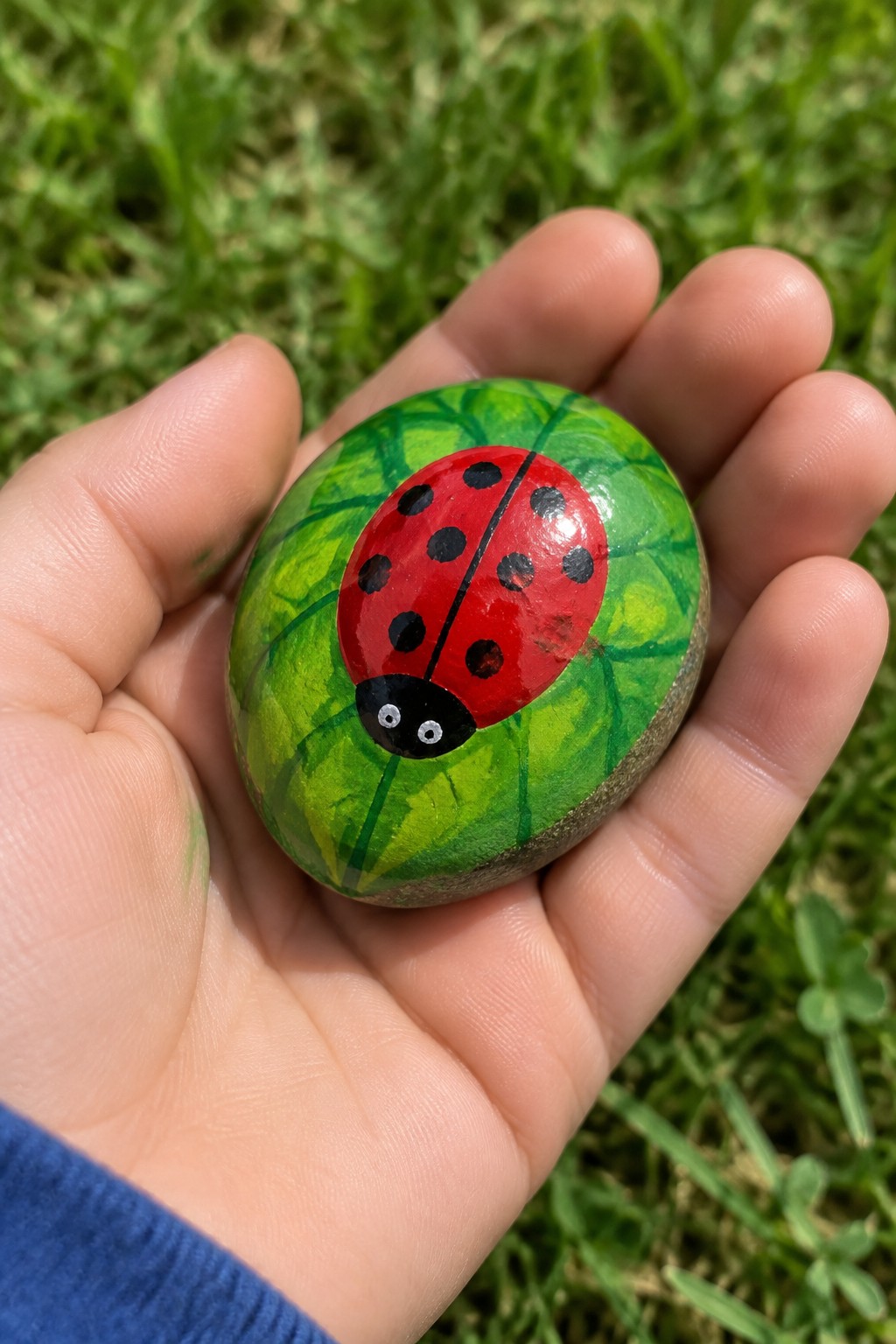 Hand holding painted rock with red ladybug on green leaves.