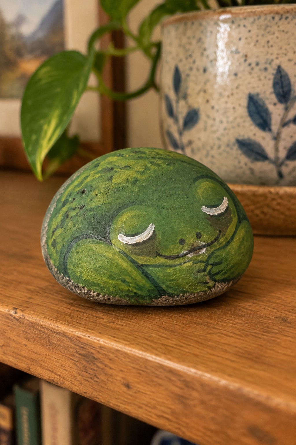 Green frog painted on smooth stone with closed eyes on wooden shelf near potted plant.