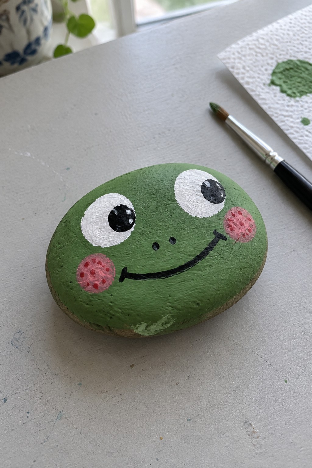 Green painted rock frog with white eyes, pink cheeks, black smile beside paintbrush